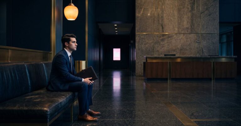Jeune candidat en costume attendant seul sur une banquette en cuir dans le hall sombre d'un siège social, illustration de l'écart entre promesse employeur et vécu candidat